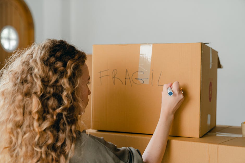 A woman with long, curly brown hair is inside a home, writing the word 'FRAGILE' in large letters on a cardboard box with a blue marker. The box is placed on top of other packed boxes in a room with a