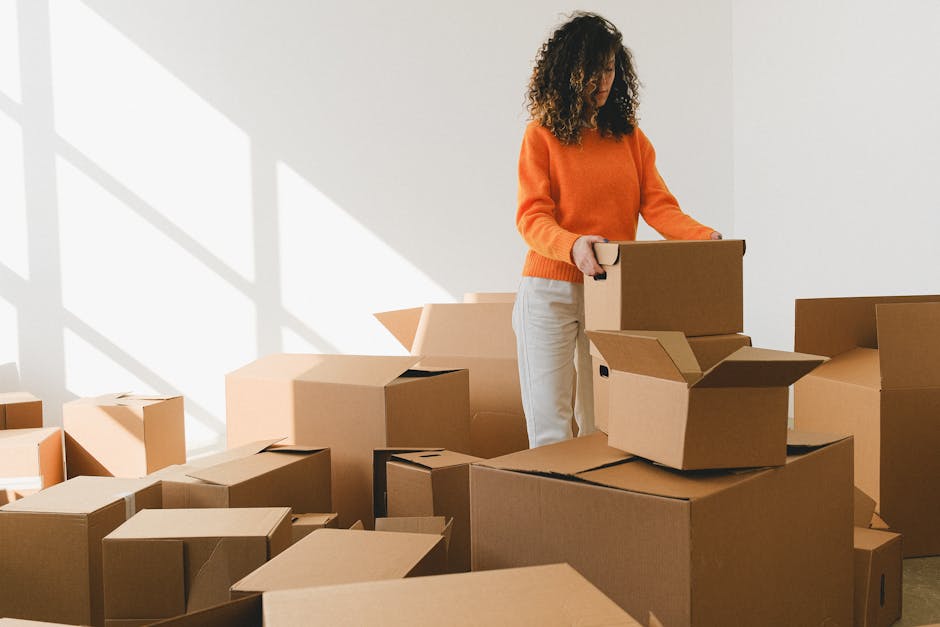 A woman with long, curly brown hair is inside a home, writing the word 'FRAGILE' in large letters on a cardboard box with a blue marker. The box is placed on top of other packed boxes in a room with a