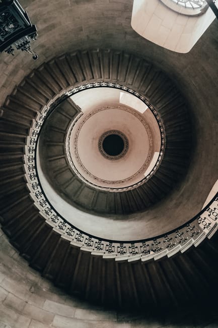 A spiraling view of a historic, narrow Victorian staircase seen from above, showing ornate white balustrades and dark wooden steps that wind downward around a central open space. The staircase is situ