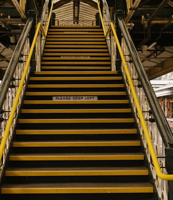 A spiraling view of a historic, narrow Victorian staircase seen from above, showing ornate white balustrades and dark wooden steps that wind downward around a central open space. The staircase is situ
